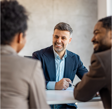Two men and one woman dressed in business attire sitting at a desk having a conversation with smiles on their faces.