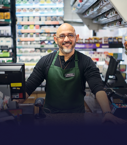 A cashier at a convenience store stands smiling behind the counter