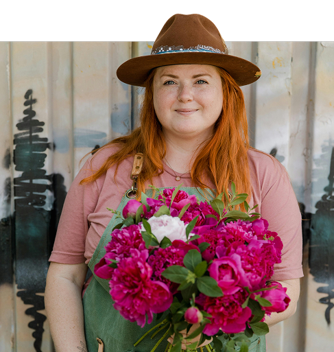 A woman in a large brown hat and an apron holds a large bouquet of pink and white flowers