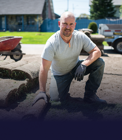 A man kneeling down in a dirt yard with a roll of sod in his hand and a pile of sod just behind him