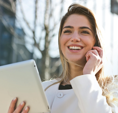 A smiling woman in an outdoor setting holds a tablet in her right hand and touches her face with her left.